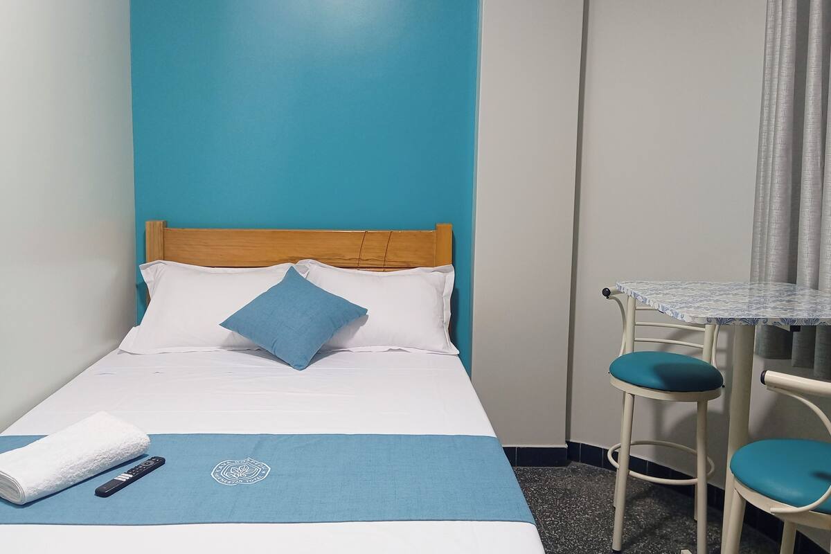 Habitación de hotel con cama, una mesita con sillas, un control remoto y una toalla sobre la cama.