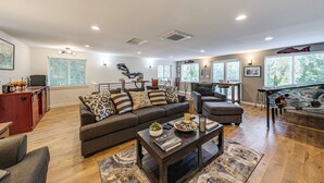 A spacious living room with a large sofa, a coffee table, and a fireplace. There is a dining area with a table and chairs, and a kitchen area with cabinets and appliances.