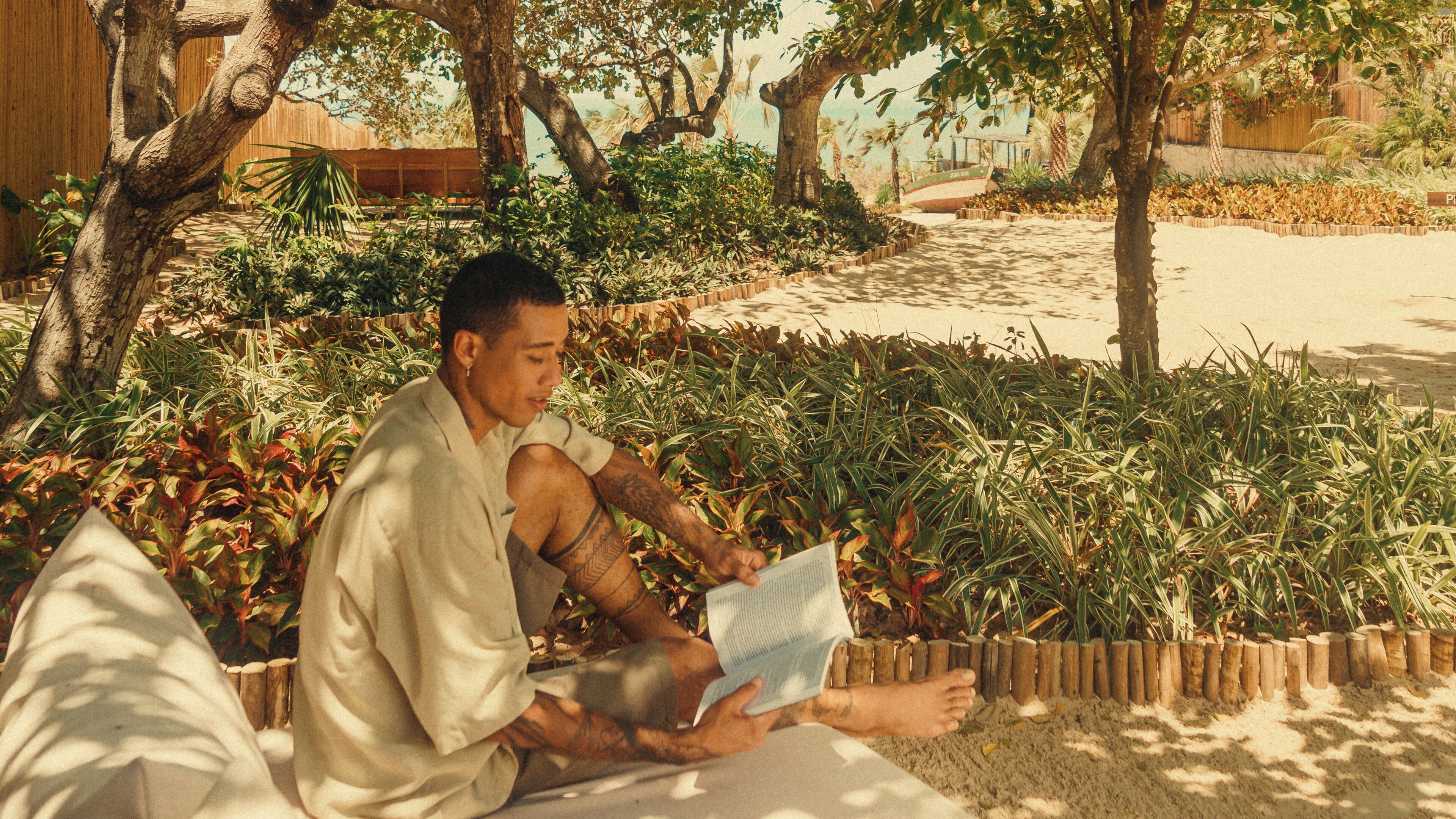 A person sitting on the ground reading a book in a garden with trees and plants.