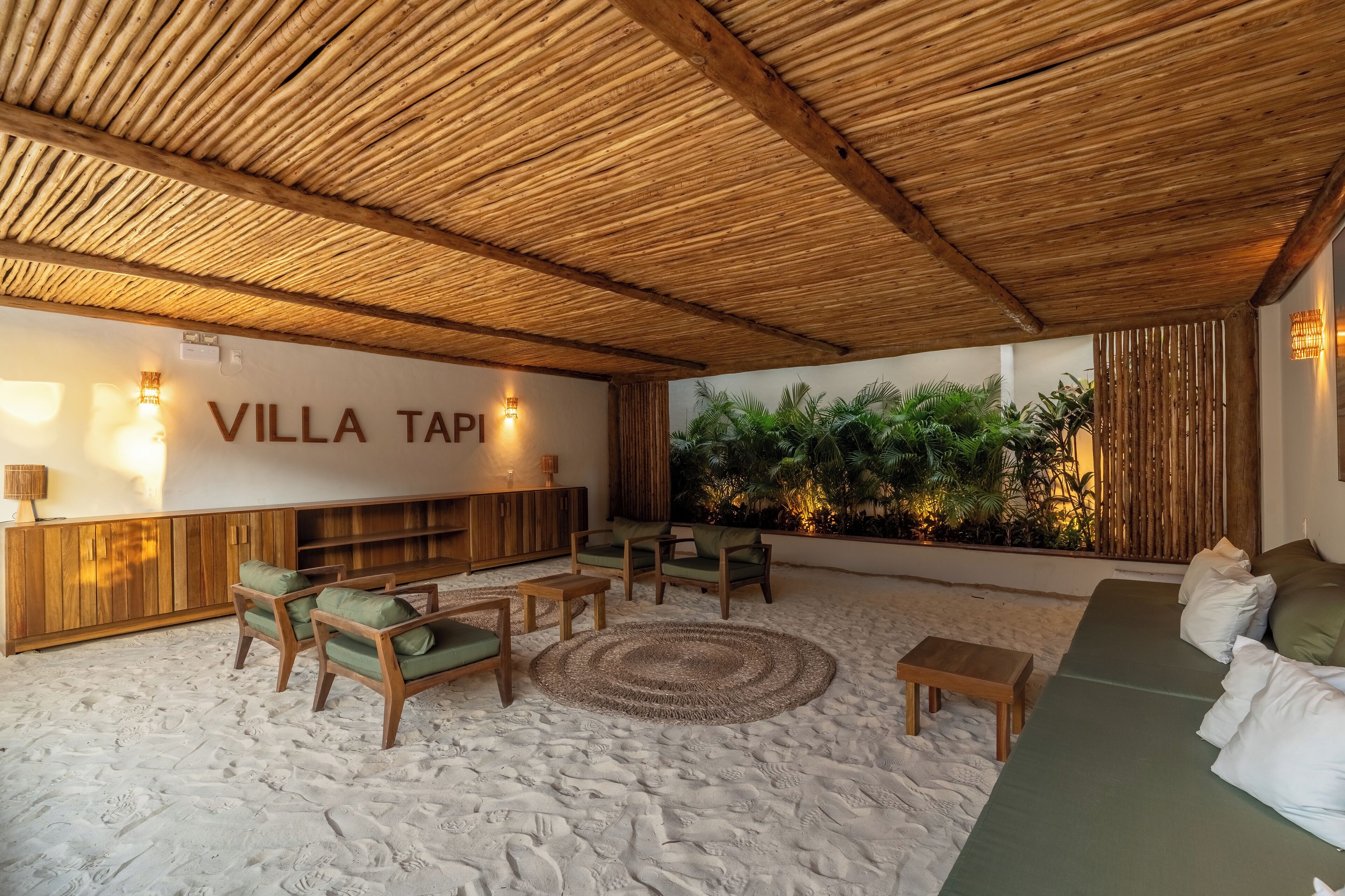 A cozy lounge area with a sandy floor, wooden furniture, and a wall with 'VILLA TAPI' signage.