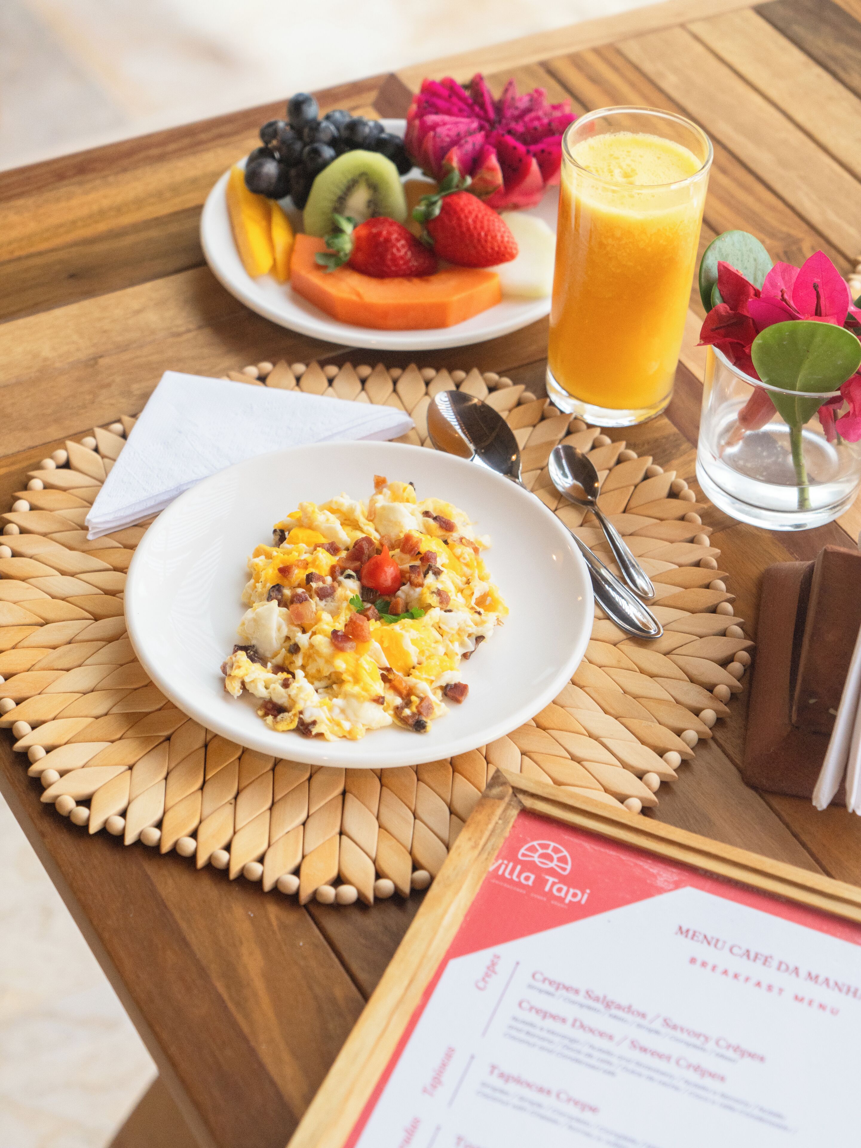 Desayuno con huevos revueltos, una bandeja de fruta, un vaso de jugo de naranja y un menú sobre una mesa de madera.