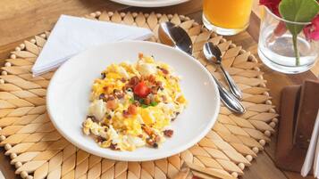 Desayuno con huevos revueltos, una bandeja de fruta, un vaso de jugo de naranja y un menú sobre una mesa de madera.