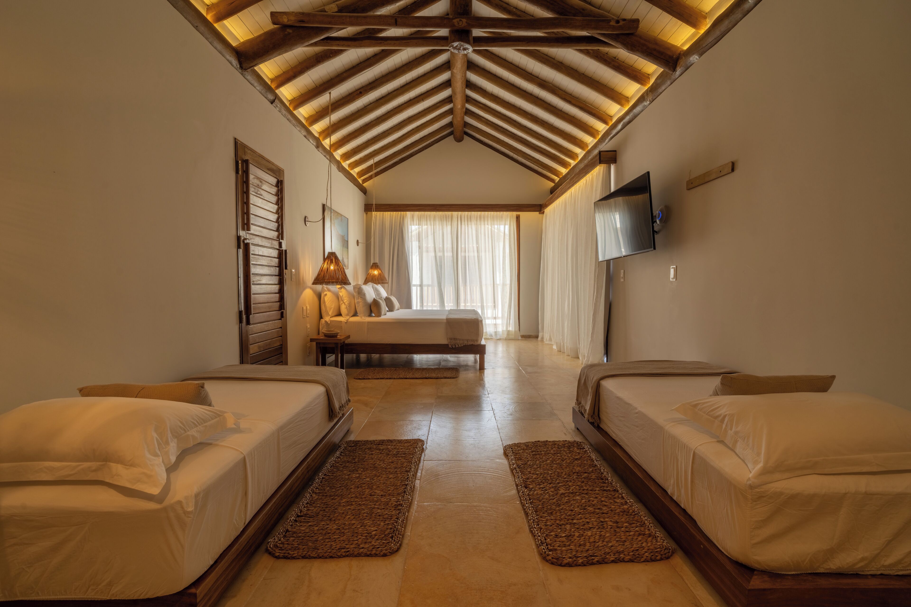 A hotel room with a high ceiling, a large bed, two smaller beds, a TV, and a window with curtains.