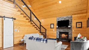 A cozy log cabin interior with a fireplace, a sofa, a chair, and a staircase. - Gettin' By Cozy 2 Bedroom Cabin with Hot Tub & WiFi in Charming Broken Bow (Broken Bow)