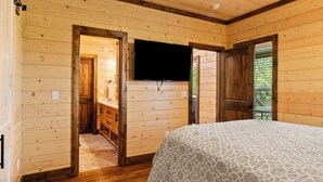 A wooden-paneled room with a bed, a television mounted on the wall, and a door leading to a balcony. - Gettin' By Cozy 2 Bedroom Cabin with Hot Tub & WiFi in Charming Broken Bow (Broken Bow)
