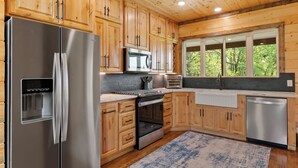 A modern kitchen with wooden cabinets, stainless steel appliances, and a large window providing a view of the outdoors. - Gettin' By Cozy 2 Bedroom Cabin with Hot Tub & WiFi in Charming Broken Bow (Broken Bow)