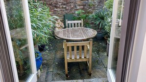 Un patio con una mesa y sillas de madera, rodeado de plantas en macetas y un muro de piedra.