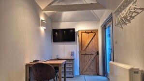 Una habitación pequeña y acogedora con un escritorio de madera, una silla, un televisor fijado a la pared y una puerta grande de madera.