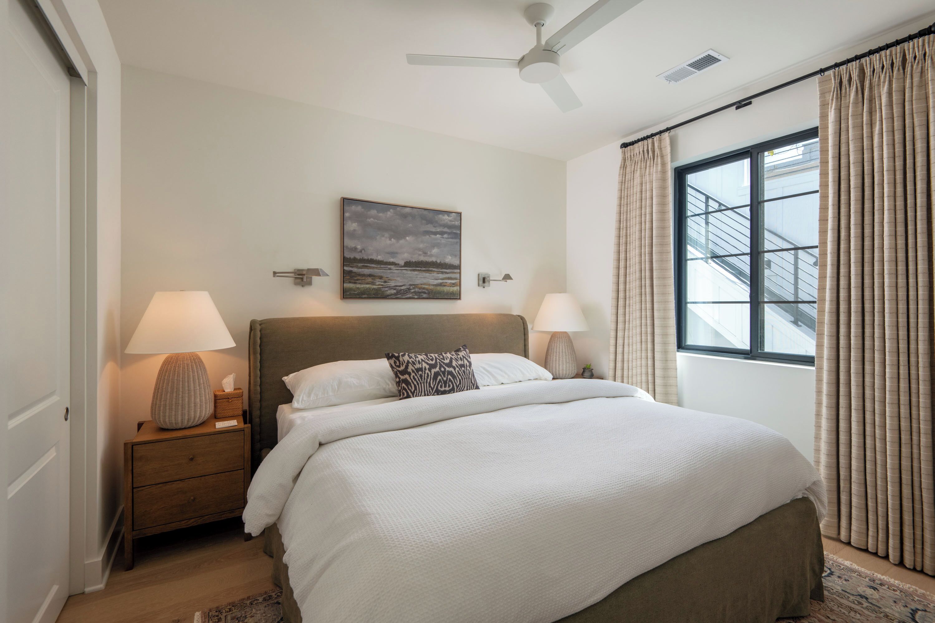 A bedroom with a bed, nightstands, a ceiling fan, a window with curtains, and a painting on the wall.