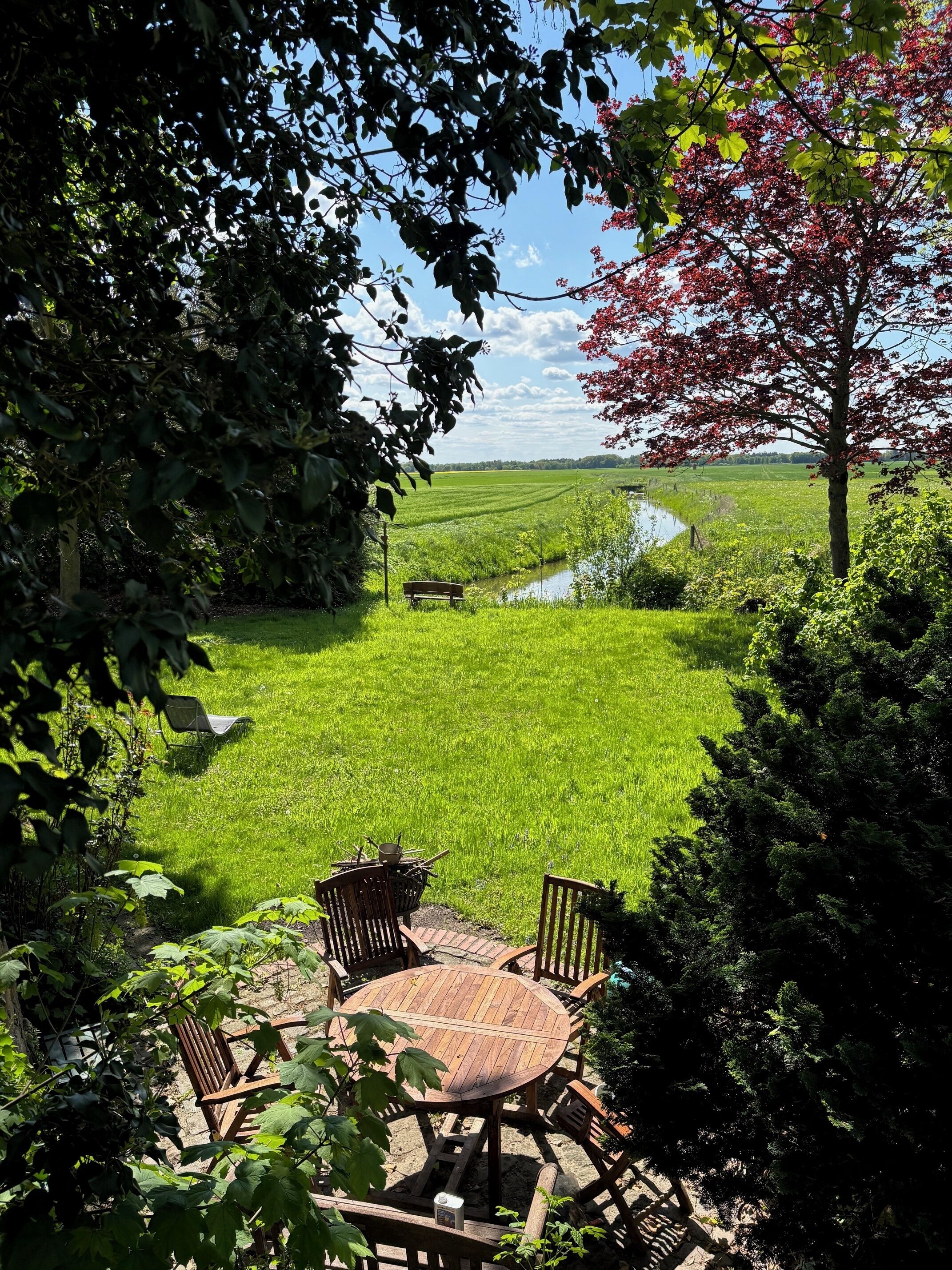 Un jardin avec une table et des chaises en bois, entouré d’une végétation luxuriante et offrant une vue sur un champ et un petit plan d’eau.