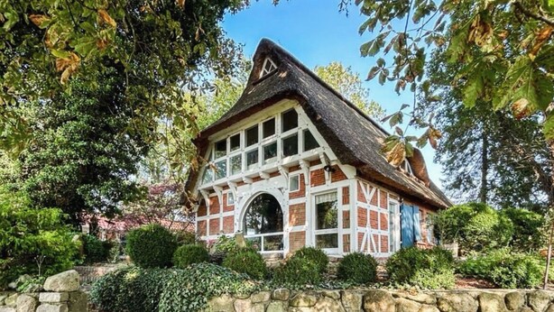 Une maison traditionnelle avec un toit à forte pente, un mur de pierre à l’avant et un petit jardin agrémenté d’arbustes et d’arbres.