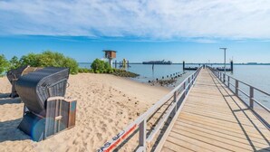 Une jetée en bois s’étendant dans des eaux calmes, avec une plage d’un côté et une petite construction de l’autre.