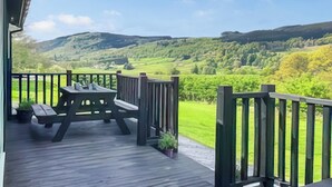 A wooden deck with picnic tables and potted plants, overlooking a green valley and hills.