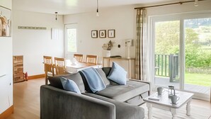 A modern living room with a grey sofa, wooden flooring, a dining area with chairs, and a view of greenery through large windows.