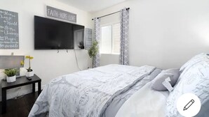 A bedroom with a bed, a TV mounted on the wall, a small table with a plant, and a window with curtains. - Sf City Ocean View. Free parking inside the compound. 15 min beach Drive (Oakland)