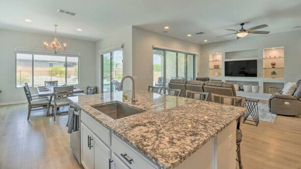 A spacious kitchen with granite countertops, a dining area with a table and chairs, a living room with a sofa and TV, and a ceiling fan. - The Desert Jewel in Lucero w/ Private Pool, Jacuzzi, Pool Table! (Goodyear)
