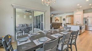 A dining area with a large table set for six, a chandelier, and a view into a living room with a sofa and a coffee table. - The Desert Jewel in Lucero w/ Private Pool, Jacuzzi, Pool Table! (Goodyear)