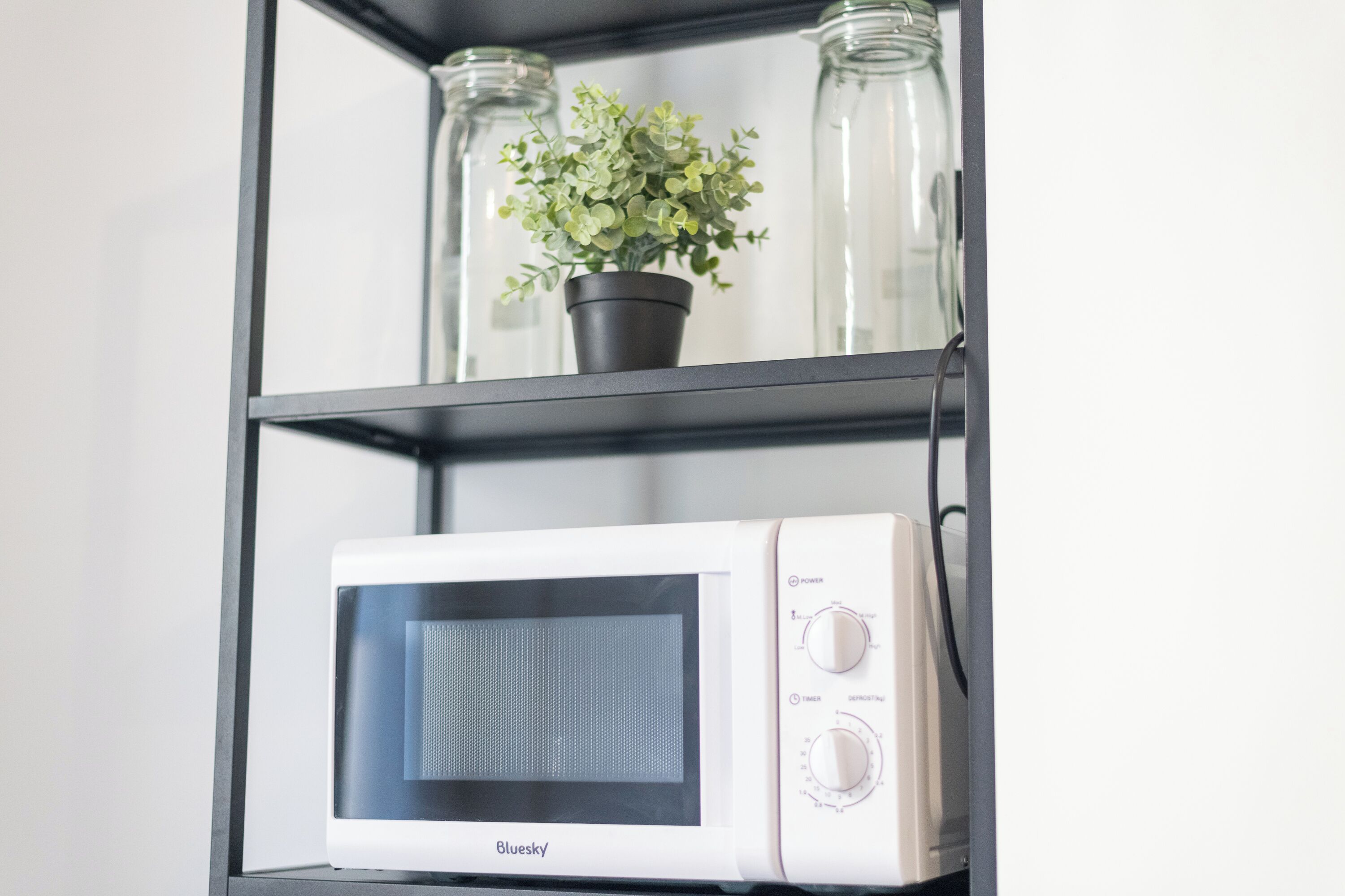 Un four à micro-ondes blanc, posé sur une étagère noire, avec une plante en pot et deux bocaux en verre.