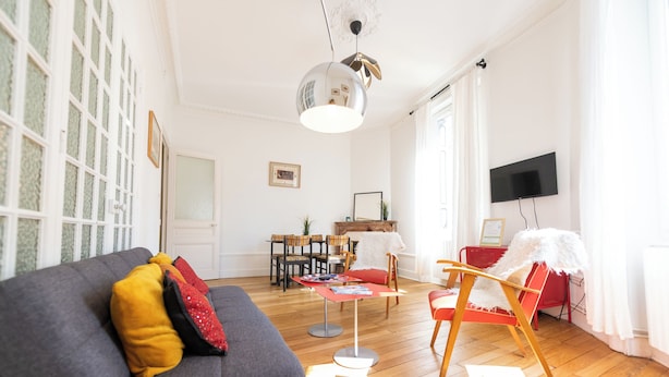A living room with a grey sofa, red and yellow cushions, a round table, and a television mounted on the wall.