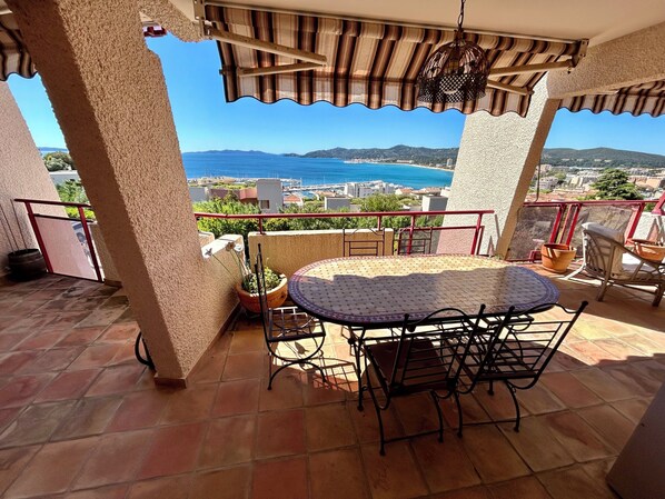 A balcony with a tiled floor, a round table with chairs, a potted plant, and a view of the sea and distant buildings. - Spacious & Unique Sea View Apt – Terrace, Pool & Charm (Le Lavandou)