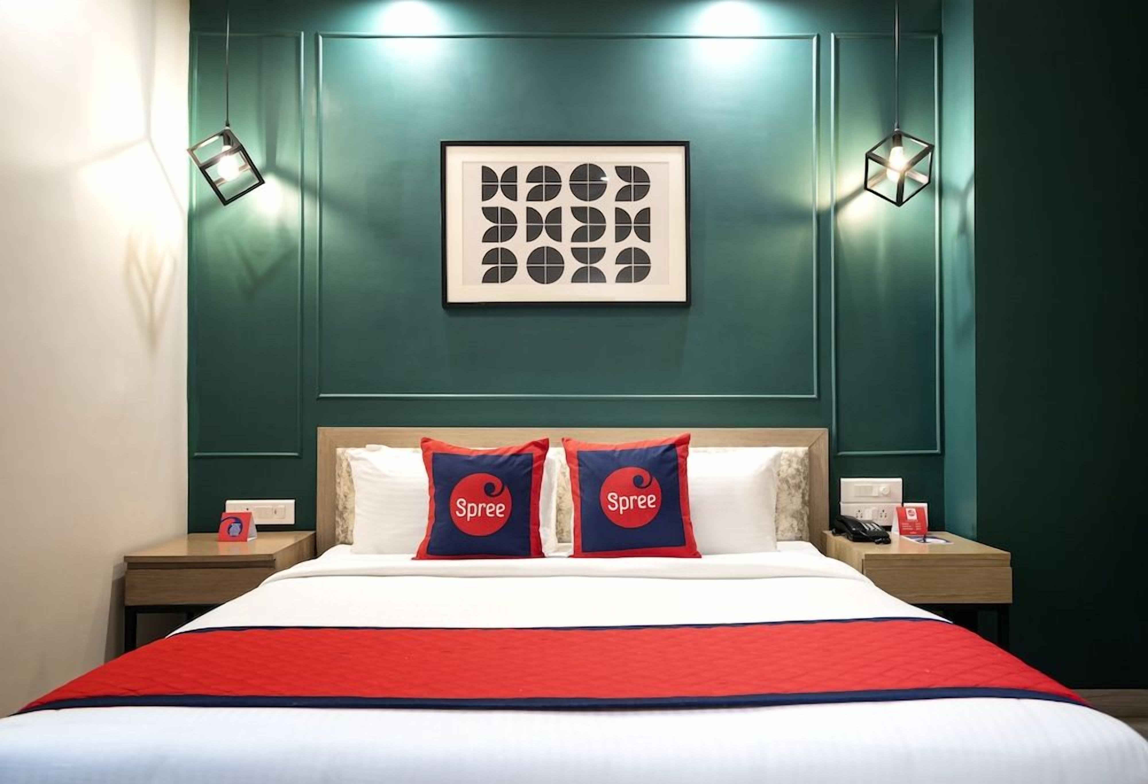 A hotel room with a bed, two bedside tables, a nightstand, a framed artwork on the wall, and two wall-mounted lamps.