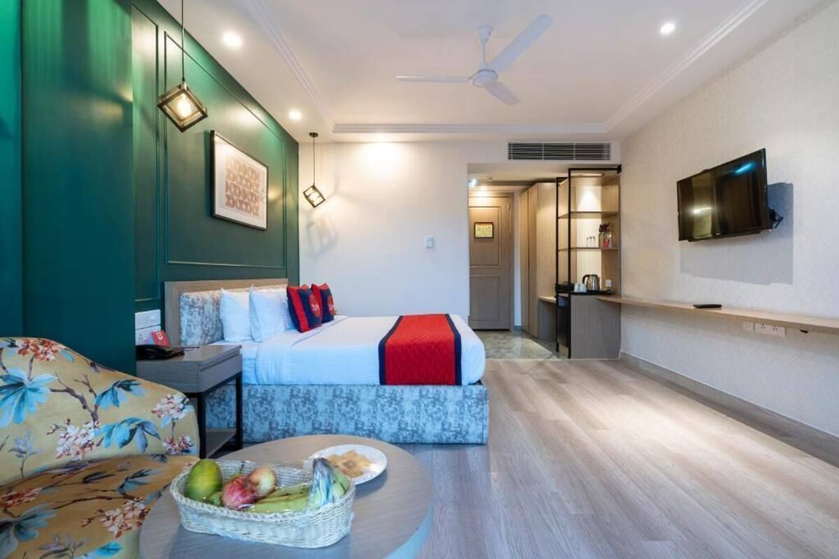 A modern hotel room with a bed, a sofa, a dining table with fruit, a TV, and a ceiling fan.