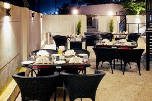 Un espace extérieur aménagé pour les repas, avec des chaises et des tables en osier, équipé de luminaires et d’une poubelle.