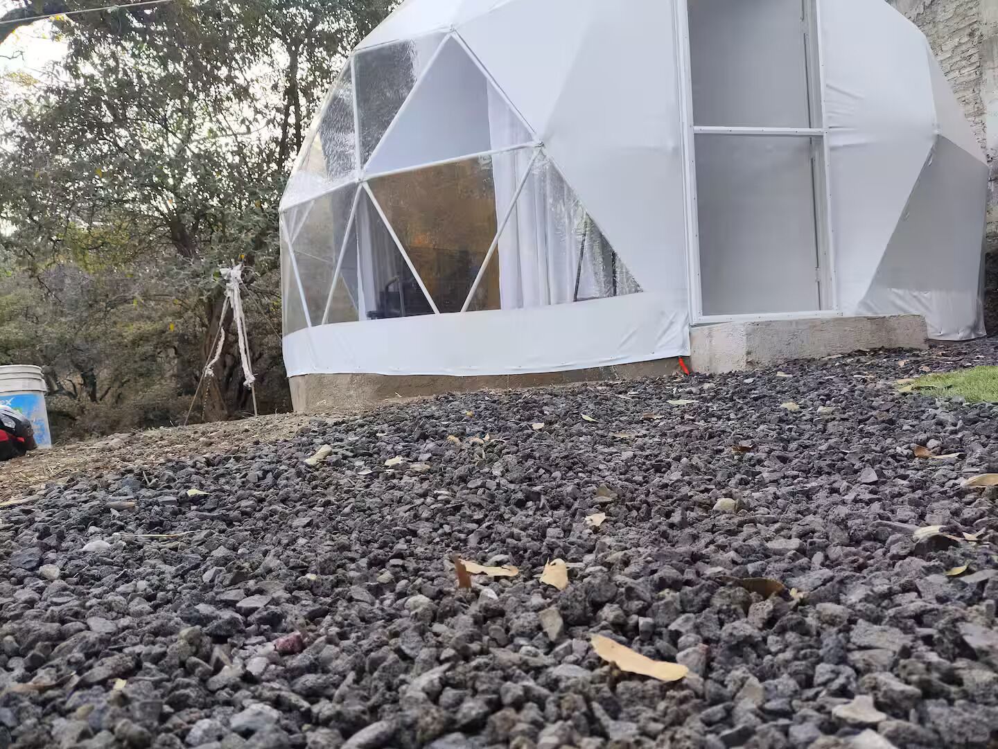 Uma estrutura em forma de cúpula geodésica com exterior branco, rodeada por brita e árvores.