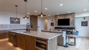 A modern kitchen with wooden cabinets, a large island, and a built-in oven. There is a stone fireplace with a flat-screen TV above it, and a cozy seating area with a sofa and a small round table. - Middle Teton 2N | Stunning Views + Private Balcony (Driggs)