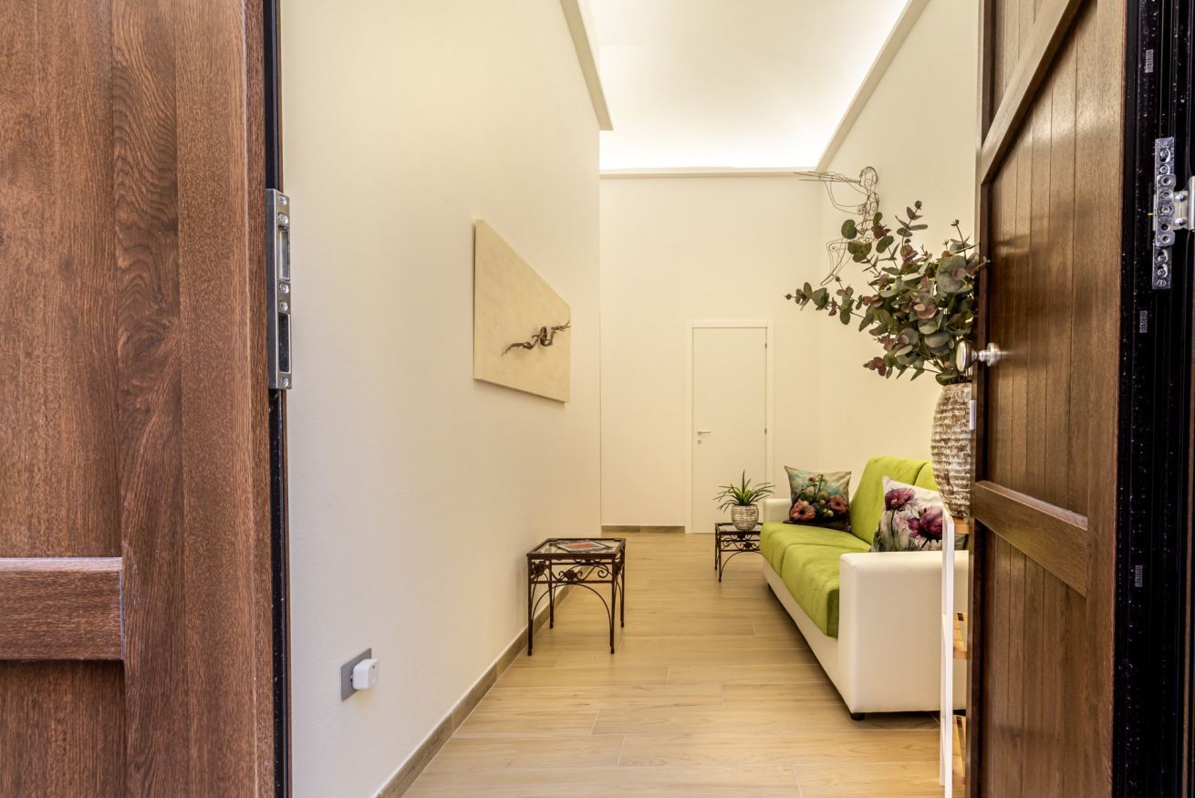 Un pasillo con una puerta de madera, un sofá blanco con cojines verdes, una mesita y una obra de arte colgada en la pared.