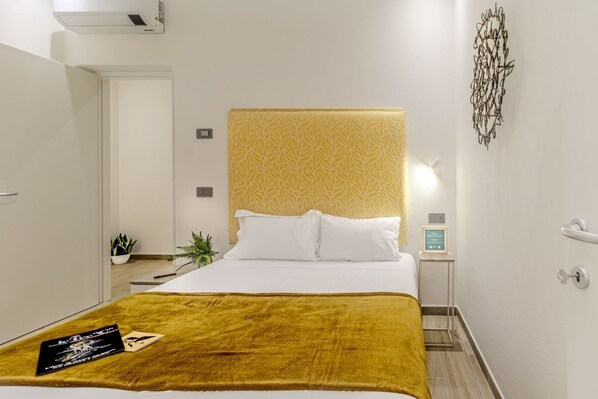 A hotel room with a bed, a bedside table, a nightstand, a wall-mounted artwork, and a door. - Artis Rooms - Suite Yellow (Cagliari)