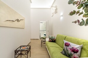 A hallway with a green sofa, a small table with a magazine, and a wall with a painting and a wall-mounted plant. - Artis Rooms - Suite Yellow (Cagliari)