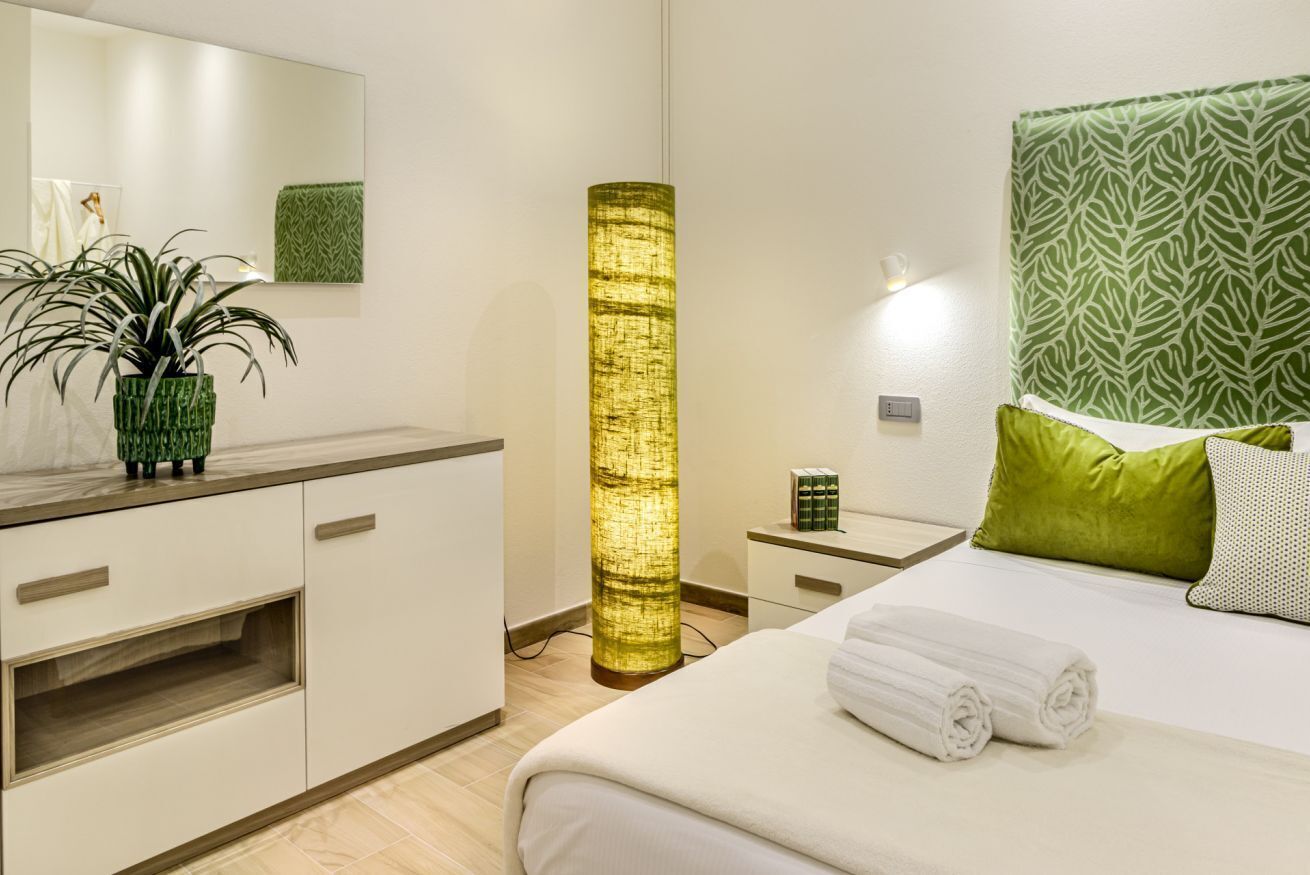 Une chambre d’hôtel comprenant un lit, une table de chevet, une petite table d’appoint, une lampe, un miroir, une plante verte et une tête de lit verte à motifs.