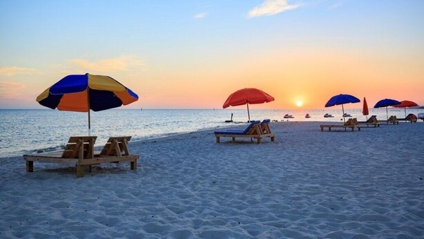 A beach with wooden lounge chairs and colorful umbrellas, a sunset in the background, and calm sea.