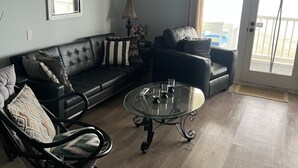 A living room with a sectional sofa, a glass coffee table, and a black armchair. - OCEANFRONT PENTHOUSE- PRIVATE BEACH ACCESS & STUNNING VIEWS – WALK TO BOARDWALK! (Carolina Beach)