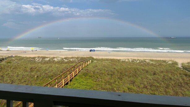 A beach with a wooden boardwalk leading to the shore, a rainbow in the sky, and the ocean in the background. - OCEANFRONT PENTHOUSE- PRIVATE BEACH ACCESS & STUNNING VIEWS – WALK TO BOARDWALK! (Carolina Beach)