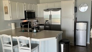 A modern kitchen with white cabinetry, stainless steel appliances, a central island with chairs, and a trash can. - OCEANFRONT PENTHOUSE- PRIVATE BEACH ACCESS & STUNNING VIEWS – WALK TO BOARDWALK! (Carolina Beach)