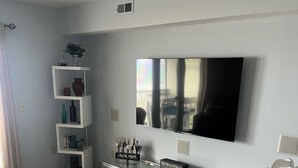 A modern living room with a flat-screen TV mounted on the wall, a white entertainment unit with electronic devices, a dining table with chairs, and a wooden floor. - OCEANFRONT PENTHOUSE- PRIVATE BEACH ACCESS & STUNNING VIEWS – WALK TO BOARDWALK! (Carolina Beach)
