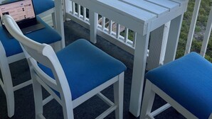 A balcony with a white table and two blue chairs, overlooking a beach with a wooden walkway and a fence. - OCEANFRONT PENTHOUSE- PRIVATE BEACH ACCESS & STUNNING VIEWS – WALK TO BOARDWALK! (Carolina Beach)