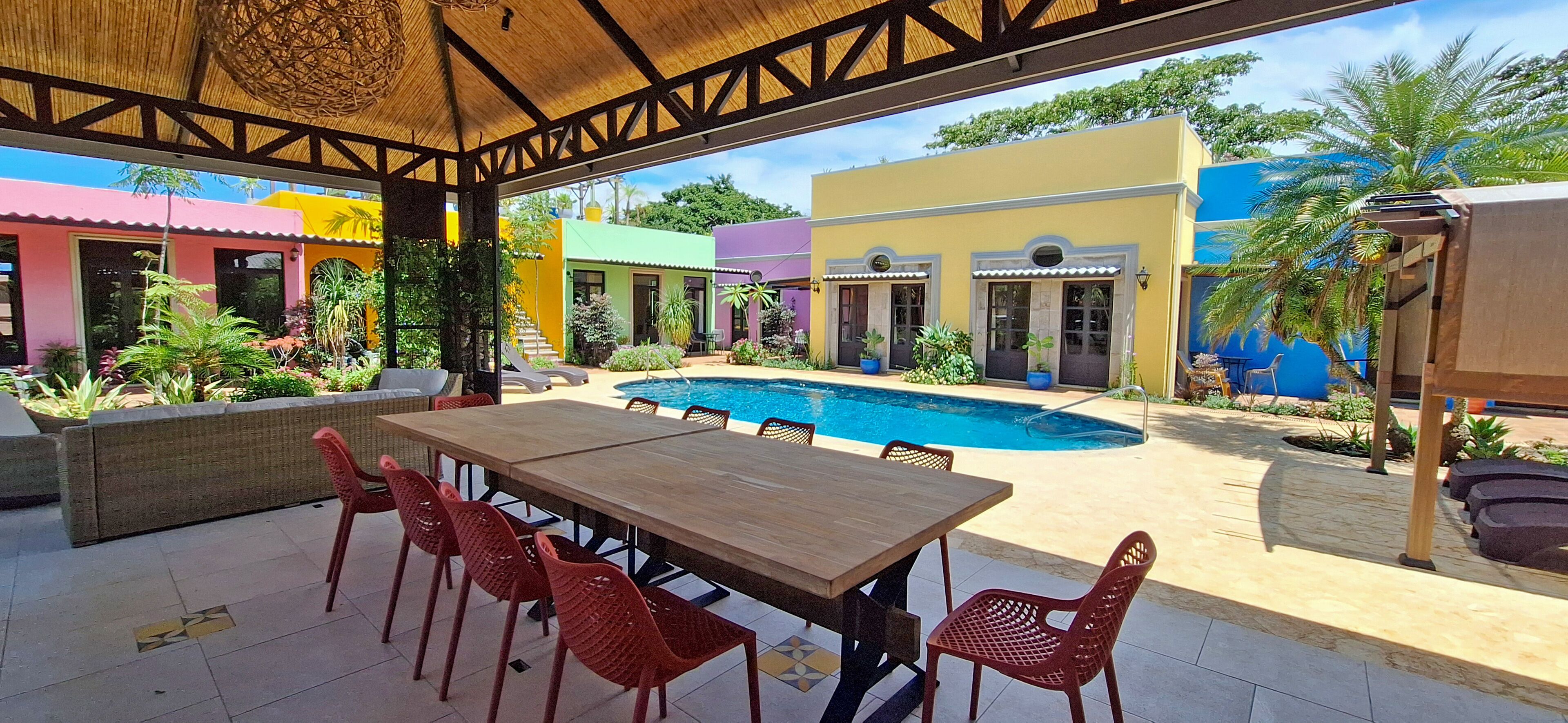 Un espacio al aire libre techado con piscina, mesa de madera y sillas rojas.