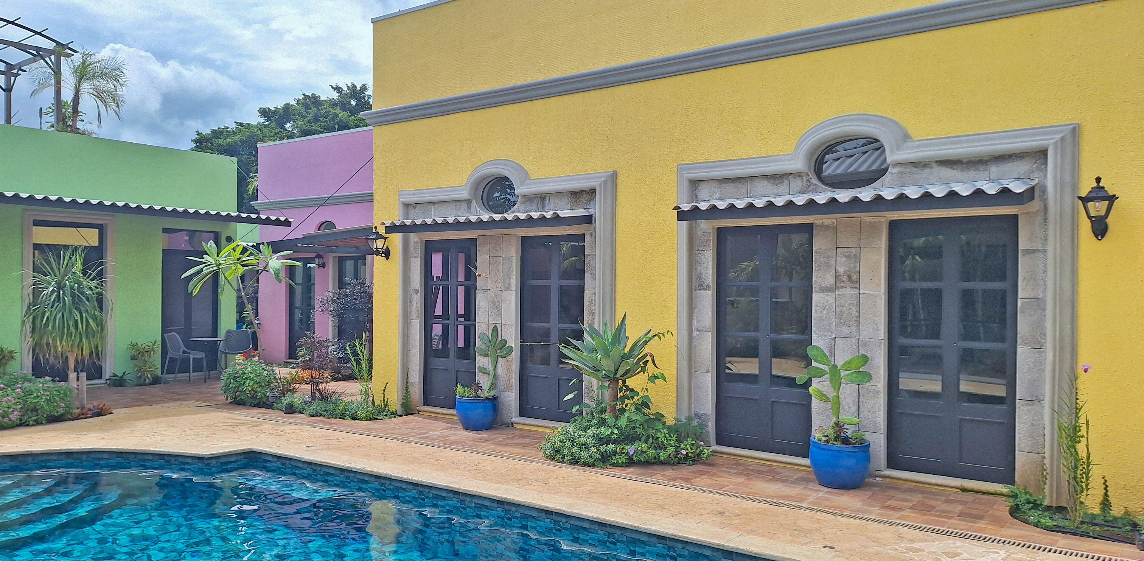 Zona junto a la piscina con un edificio amarillo, uno verde y otro rosa. Hay plantas en macetas y una piscina.