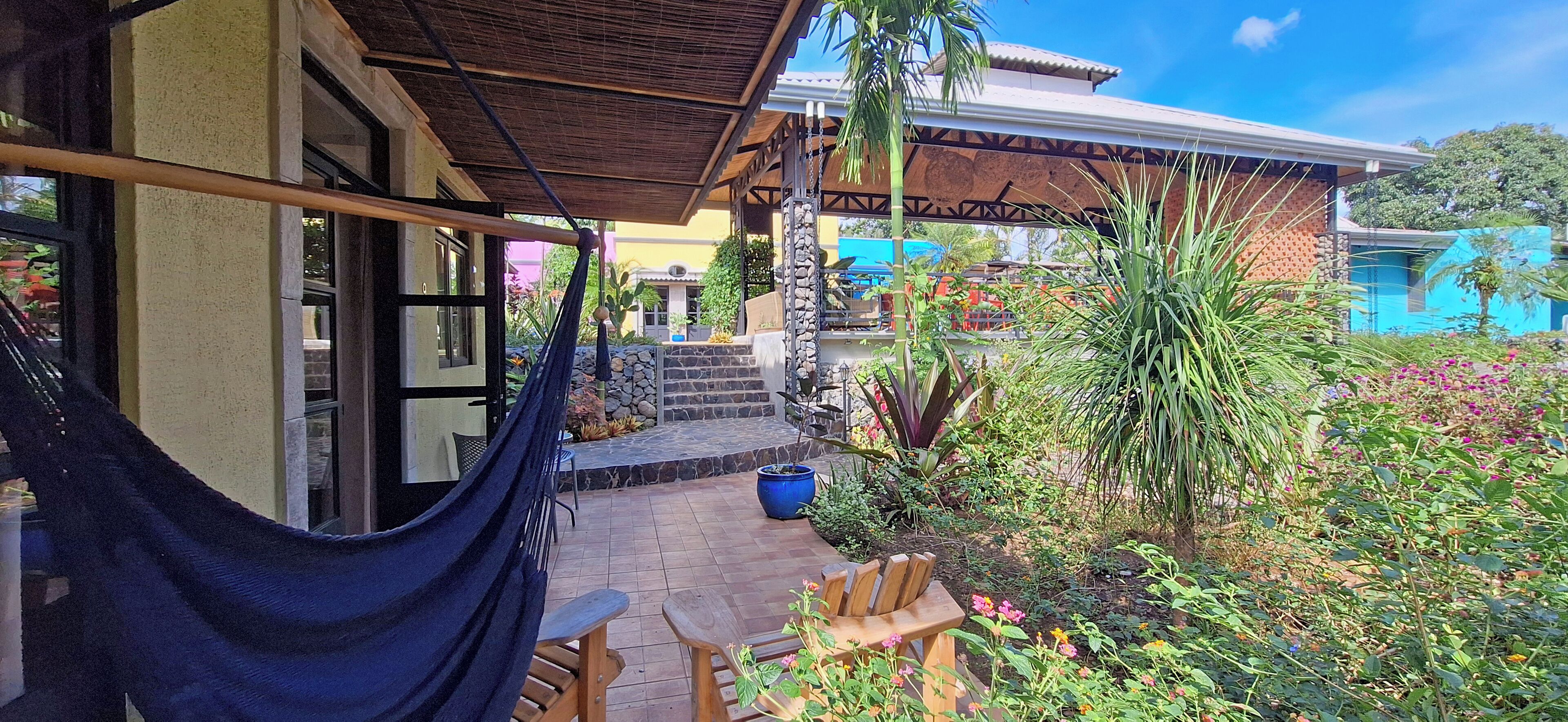 A covered outdoor seating area with a hammock, wooden chairs, and potted plants.