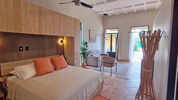 A bedroom with a wooden headboard, a bed with white and orange pillows, a ceiling fan, and a view of the patio area with a chair and a plant.