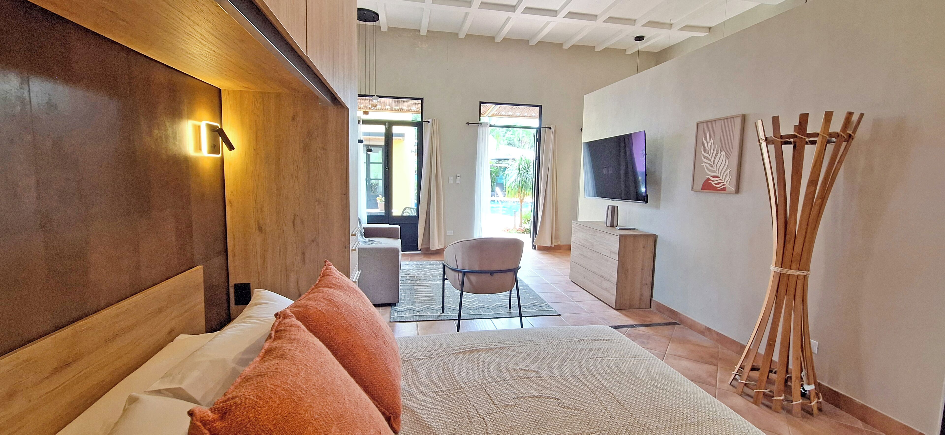 A modern hotel room with a wooden headboard, a large bed with orange pillows, a flat-screen TV, a wooden sculpture, and a view of the outdoor area through a glass door.