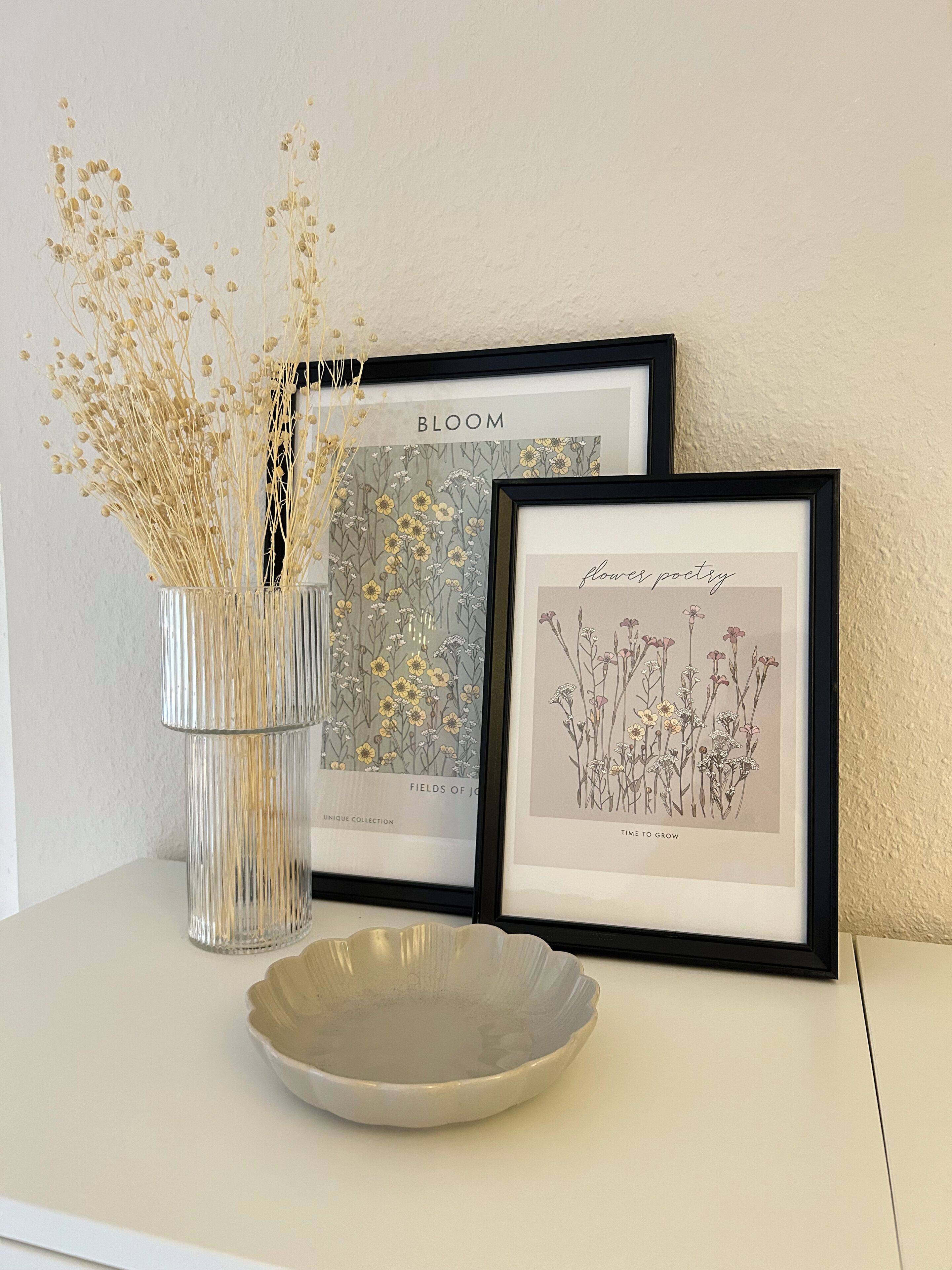 A framed botanical print, a glass vase with dried plants, and a ceramic bowl on a white table against a textured wall.