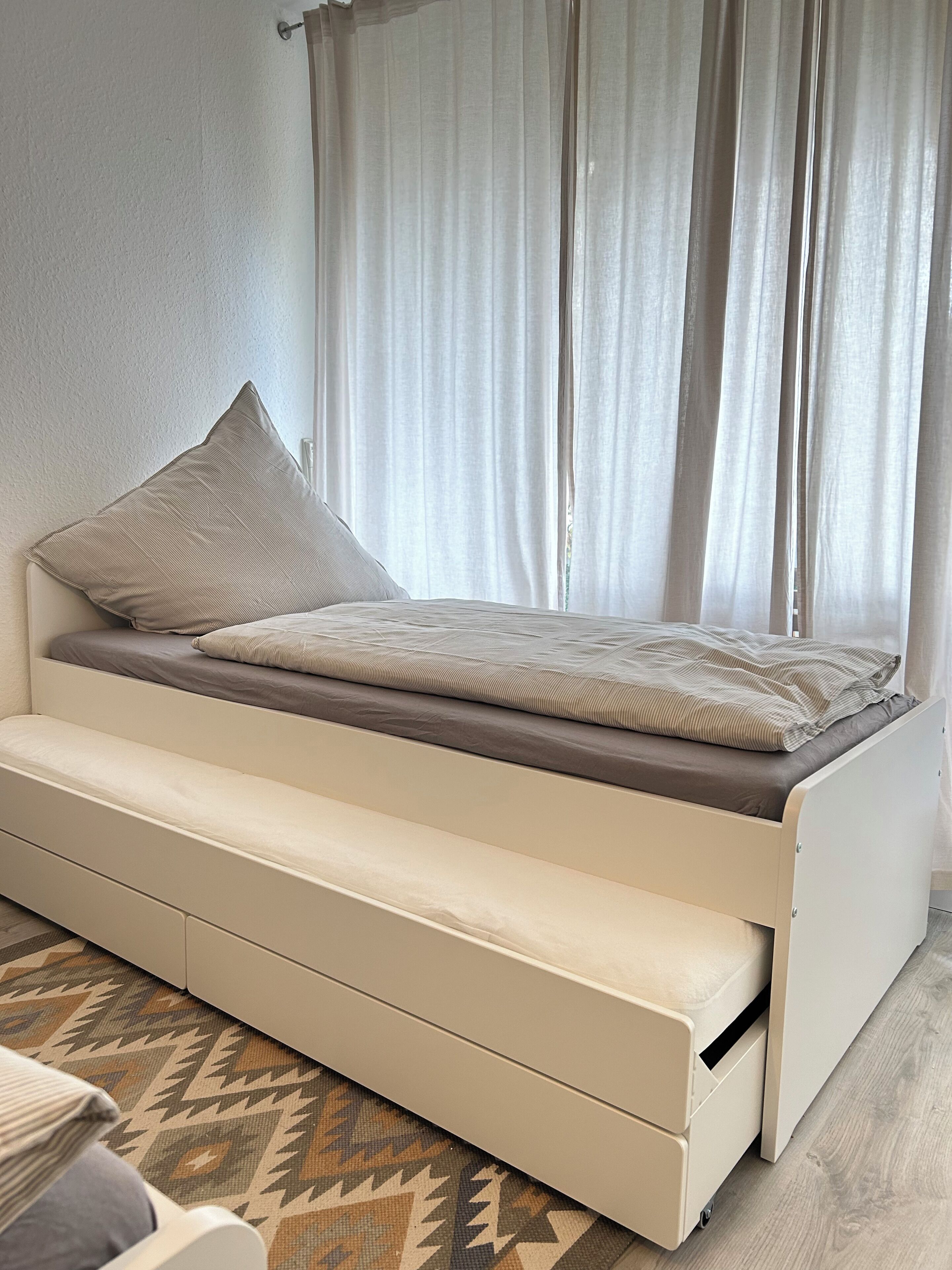 Ein platzsparendes Bett mit ausziehbarer Schublade, ein Fenster mit durchsichtigen Vorhängen und ein gemustertes Teppich.