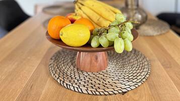 A wooden table with a woven placemat, a fruit bowl containing oranges, lemons, bananas, and grapes, and a hanging lamp.
