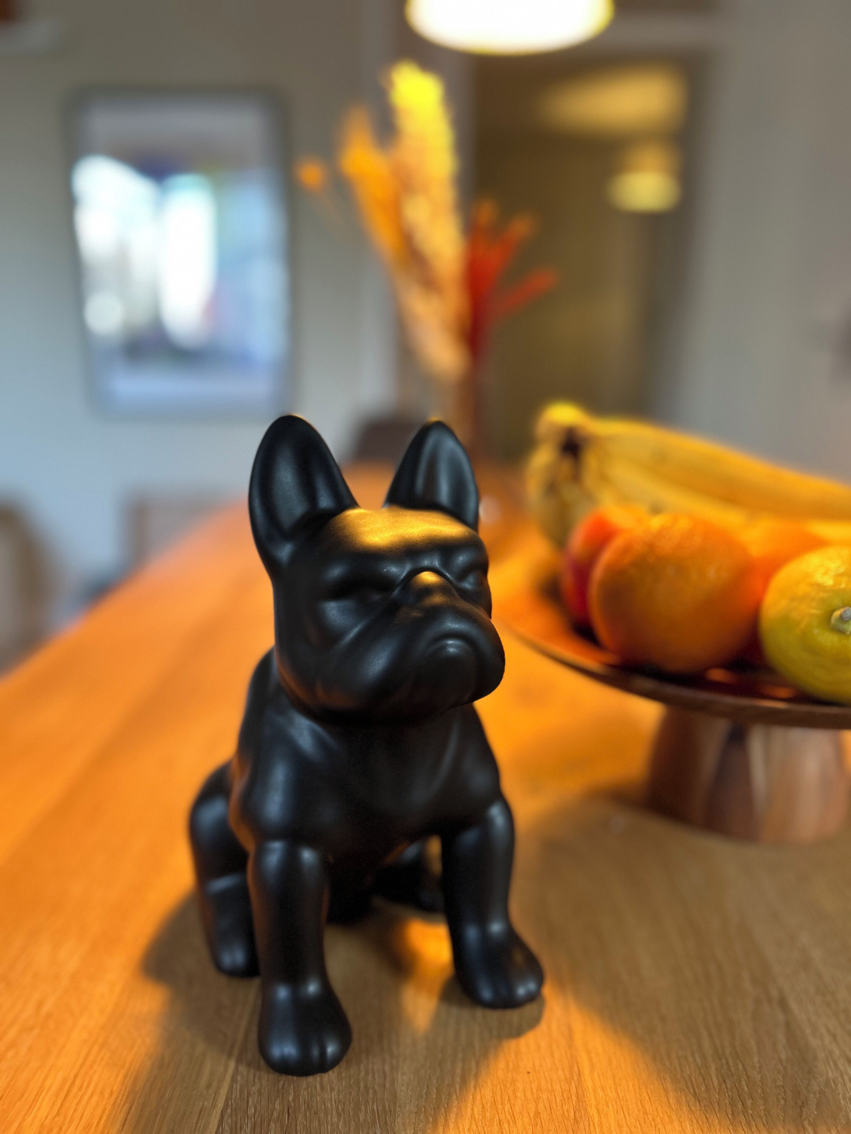 A black dog figurine on a wooden table with a fruit bowl in the background.
