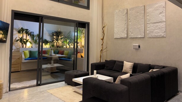 Een moderne woonkamer met een zwarte bank, een glazen deur naar een balkon en uitzicht op palmbomen.
