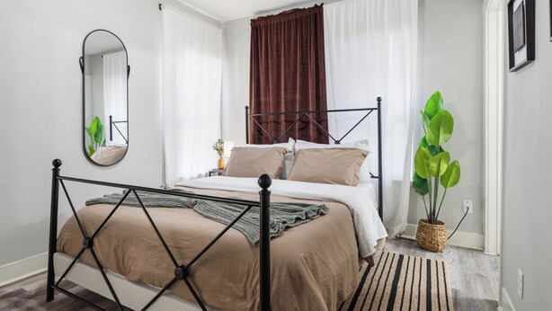 Un dormitorio con una cama, un espejo, cortinas, una planta en maceta y una alfombra.